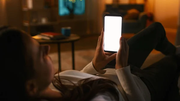 Noche en casa: Mujer joven Descansando en un sofá usando con un teléfono inteligente blanco de pantalla falsa. Chica Usando Chroma Key Teléfono Móvil, Navegación por Internet, Publicar en las redes sociales. — Foto de Stock