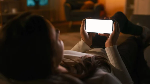 自宅での夜:若い女性ホワイトモックアップ画面スマートフォンで使用してソファの上で休憩。風景の中にクロマキー携帯電話を使用して女の子水平モード,インターネットブラウジング,ソーシャルネットワークに投稿 — ストック写真