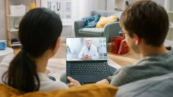 Giovane coppia di una ragazza e l'uomo seduto a casa, Utilizzando computer portatile per parlare con un medico tramite Video Conference Medical App. Hanno conversazione con il medico utilizzando Online Video Chat Applicazione. — Foto Stock