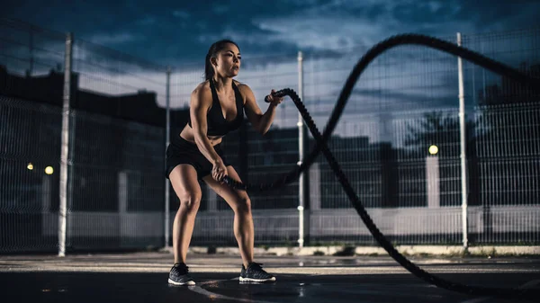 Schöne energiegeladene Fitness-Mädchen, die Übungen mit Kampfseilen machen. Sie macht ein Workout auf einem eingezäunten Basketballplatz im Freien. Abend nach dem Regen in einem Wohngebiet. — Stockfoto