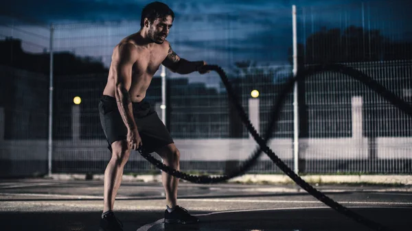 Ισχυρή μυϊκή εφαρμογή Shirtless νεαρός άνδρας κάνει ασκήσεις με σχοινιά μάχης. Κάνει μια προπόνηση σε ένα περιφραγμένο εξωτερικό γήπεδο μπάσκετ. Βράδυ μετά τη βροχή σε μια κατοικημένη περιοχή της γειτονιάς. — Φωτογραφία Αρχείου