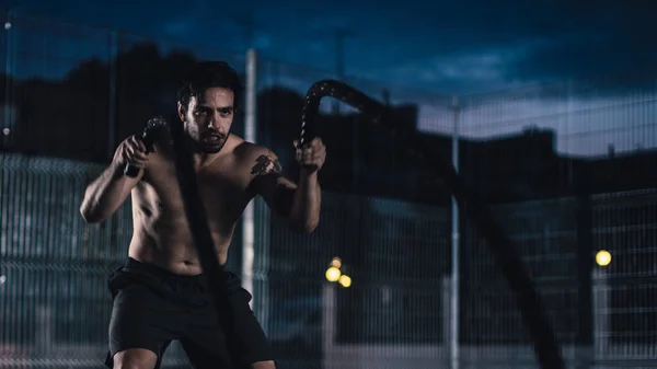 Forte Muscular Fit Shirtless Young Man está fazendo exercícios com cordas de batalha. Ele está fazendo um treino em um campo de basquete ao ar livre cercado. Noite após a chuva em uma área de vizinhança residencial. — Fotografia de Stock