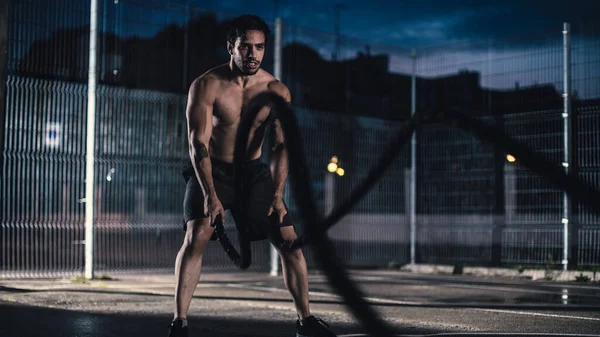 Strong Muscular Fit Shirtless Young Man está haciendo ejercicios con cuerdas de batalla. Está haciendo un entrenamiento en una cancha de baloncesto al aire libre vallada. Noche después de la lluvia en un barrio residencial. — Foto de Stock