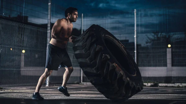 Silny muskularny Fit Młody człowiek bez koszuli robi ćwiczenia na ogrodzonym boisku do koszykówki na świeżym powietrzu. On Flipping a Big Heavy Tire w godzinach popołudniowych Po deszczu w dzielnicy mieszkalnej. — Zdjęcie stockowe