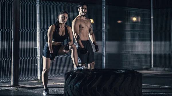 Beautiful Energetic Fitness Girl and Muscular Male Posing After Exercises in a Fenced Outdoor Basketball Court. They Were Flipping a Big Heavy Tire in a Foggy Night After Rain in a Residential