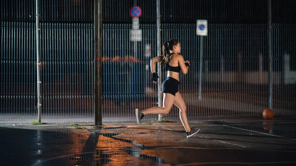 Menina Aptidão Energética bonita é Sprinting em uma quadra de basquete ao ar livre cercada. Ela corre à noite depois da chuva em uma área residencial de vizinhança. — Fotografia de Stock