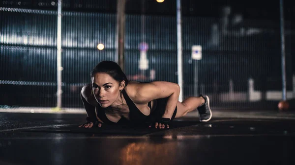 Schöne energiegeladene Fitness-Mädchen machen Liegestützübungen. Sie macht ein Workout auf einem eingezäunten Basketballplatz im Freien. Abend nach dem Regen in einem Wohngebiet. — Stockfoto