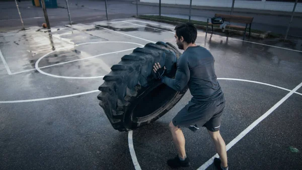 Sterke gespierde pasvorm jongeman in sportkleding en handschoenen doet oefeningen in een omheind buitenbasketbalveld. Hij flipt een zware band in een namiddag omgeving na regen. Achtergrondafbeelding — Stockfoto