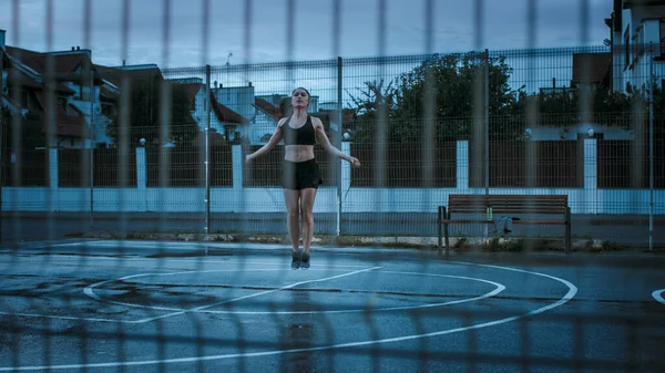 Bella Energetic Fitness Girl in Top atletico nero e pantaloncini sta saltando corda di salto. Sta facendo un allenamento in un campo da basket all'aperto recintato. Vista da dietro la recinzione. Sera dopo la pioggia. — Foto Stock