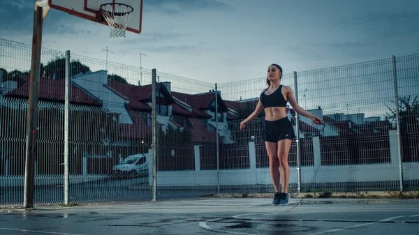 Schöne energetische Fitness-Mädchen Springseil springen. Sie macht ein Workout auf einem eingezäunten Basketballplatz im Freien. Abendschuss nach Regen in Wohngegend. — Stockfoto