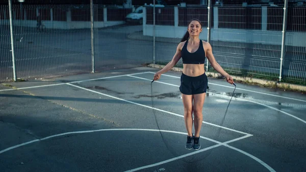 Gyönyörű mosolygós Fitness lány ugráló ugráló kötél. Edzést tart egy bekerített szabadtéri kosárlabdapályán. Esti lövés esőzés után a lakóövezetben. — Stock Fotó