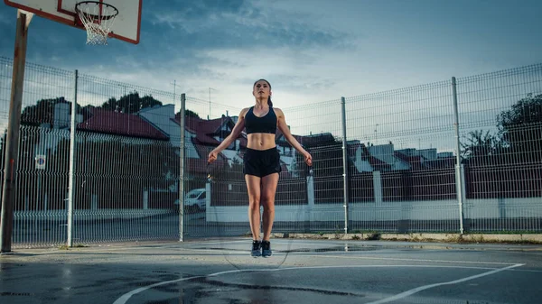 Menina Aptidão Energética bonita pulando corda de salto. Ela está fazendo um treino em uma quadra de basquete ao ar livre cercada. Disparo da noite após a chuva em uma área residencial do bairro. — Fotografia de Stock