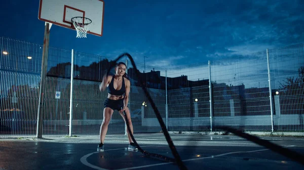 Schöne energiegeladene Fitness-Mädchen, die Übungen mit Kampfseilen machen. Sie macht ein Workout auf einem eingezäunten Basketballplatz im Freien. Abend nach dem Regen in einem Wohngebiet. — Stockfoto