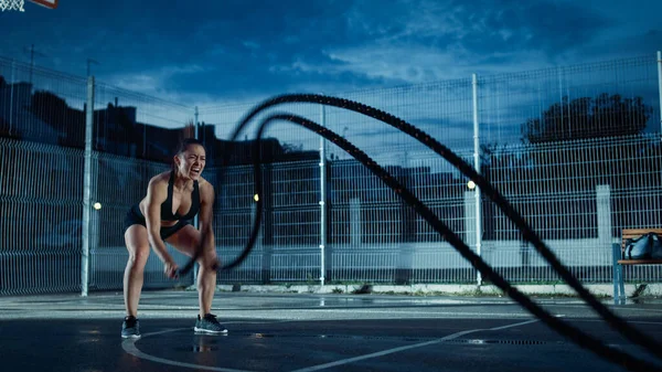 Bela aptidão energética menina fazendo exercícios com cordas de batalha. Ela está fazendo um treino em um campo de basquete ao ar livre cercado e gritando. Noite após a chuva em uma área de vizinhança residencial. — Fotografia de Stock