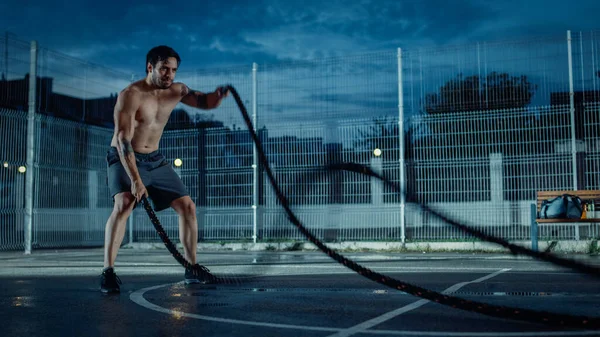 Сильный мускулистый здоровый молодой человек делает упражнения с верёвками. Он делает тренировку на закрытом баскетбольном корте. Вечер после дождя в районе жилого района. — стоковое фото
