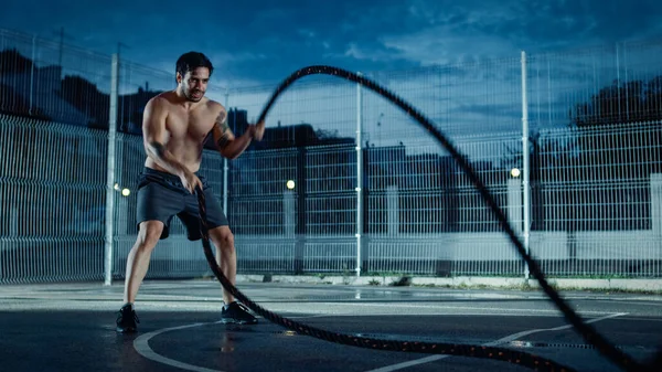 Forte Muscular Fit Shirtless Young Man está fazendo exercícios com cordas de batalha. Ele está fazendo um treino em um campo de basquete ao ar livre cercado. Noite após a chuva em uma área de vizinhança residencial. — Fotografia de Stock