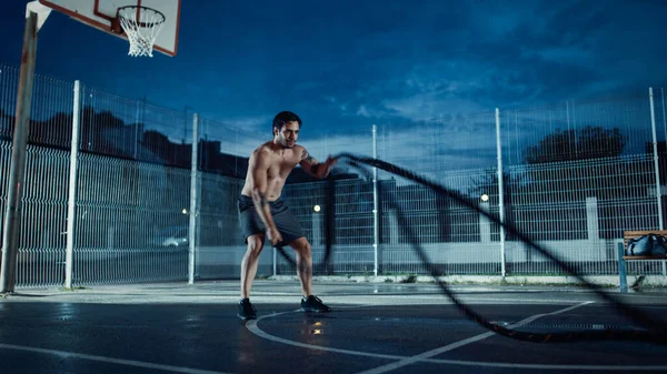 Strong Muscular Fit Shirtless Young Man is Doing Exercises with Battle Ropes. He is Doing a Workout in a Fenced Outdoor Basketball Court. Evening After Rain in a Residential Neighborhood Area. — Stock Photo, Image