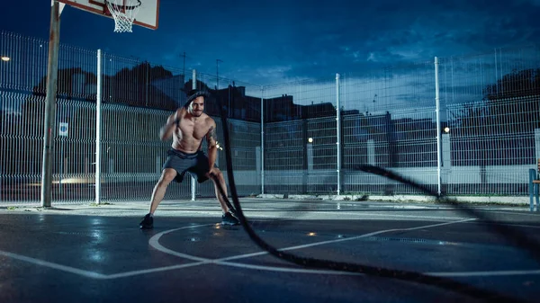 Forte Muscular Fit Shirtless Young Man está fazendo exercícios com cordas de batalha. Ele está fazendo um treino em um campo de basquete ao ar livre cercado. Noite após a chuva em uma área de vizinhança residencial. — Fotografia de Stock