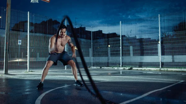 Forte Muscular Fit Shirtless Young Man está fazendo exercícios com cordas de batalha. Ele está fazendo um treino em um campo de basquete ao ar livre cercado. Noite após a chuva em uma área de vizinhança residencial. — Fotografia de Stock