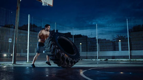 Strong Muscular Fit Young Shirtless Man κάνει ασκήσεις σε ένα περιφραγμένο εξωτερικό γήπεδο μπάσκετ. Hes Flipping ένα μεγάλο βαρύ λάστιχο σε ένα απόγευμα μετά τη βροχή σε μια κατοικημένη περιοχή της γειτονιάς. — Φωτογραφία Αρχείου