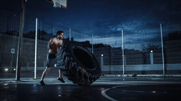 Silny muskularny Fit Młody człowiek bez koszuli robi ćwiczenia na ogrodzonym boisku do koszykówki na świeżym powietrzu. On Flipping a Big Heavy Tire w nocy po deszczu w dzielnicy mieszkalnej. — Zdjęcie stockowe