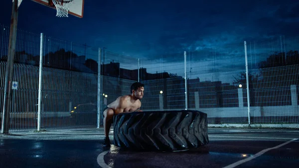 Forte Muscular Fit Young Shirtless Man está fazendo exercícios em uma quadra de basquete ao ar livre cercada. Ele está lançando um pneu pesado grande em uma tarde depois da chuva em uma área residencial de vizinhança. — Fotografia de Stock