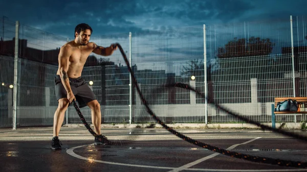 Сильный мускулистый здоровый молодой человек делает упражнения с верёвками. Он делает тренировку на закрытом баскетбольном корте. Вечер после дождя в районе жилого района. — стоковое фото
