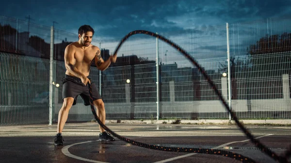 Ισχυρή μυϊκή εφαρμογή Shirtless νεαρός άνδρας κάνει ασκήσεις με σχοινιά μάχης. Κάνει μια προπόνηση σε ένα περιφραγμένο εξωτερικό γήπεδο μπάσκετ. Βράδυ μετά τη βροχή σε μια κατοικημένη περιοχή της γειτονιάς. — Φωτογραφία Αρχείου