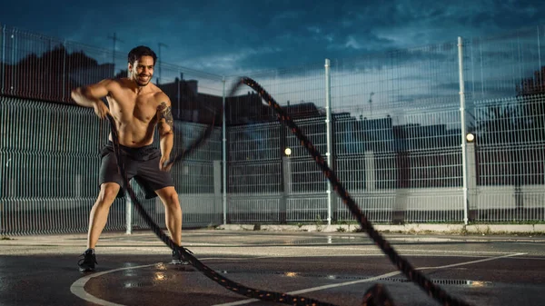 Forte Muscular Fit Shirtless Young Man está fazendo exercícios com cordas de batalha. Ele está fazendo um treino em um campo de basquete ao ar livre cercado. Noite após a chuva em uma área de vizinhança residencial. — Fotografia de Stock