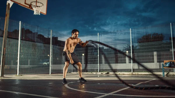 Forte Muscular Fit Shirtless Young Man está fazendo exercícios com cordas de batalha. Ele está fazendo um treino em um campo de basquete ao ar livre cercado. Noite após a chuva em uma área de vizinhança residencial. — Fotografia de Stock