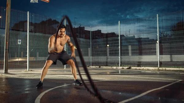 Ισχυρή μυϊκή εφαρμογή Shirtless νεαρός άνδρας κάνει ασκήσεις με σχοινιά μάχης. Κάνει μια προπόνηση σε ένα περιφραγμένο εξωτερικό γήπεδο μπάσκετ. Βράδυ μετά τη βροχή σε μια κατοικημένη περιοχή της γειτονιάς. — Φωτογραφία Αρχείου