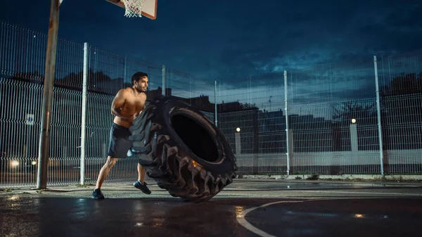 Silny muskularny Fit Młody człowiek bez koszuli robi ćwiczenia na ogrodzonym boisku do koszykówki na świeżym powietrzu. On Flipping a Big Heavy Tire w godzinach popołudniowych Po deszczu w dzielnicy mieszkalnej. — Zdjęcie stockowe