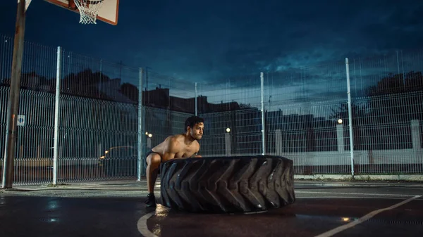 Forte Muscular Fit Young Shirtless Man está fazendo exercícios em uma quadra de basquete ao ar livre cercada. Ele está lançando um pneu pesado grande em uma tarde depois da chuva em uma área residencial de vizinhança. — Fotografia de Stock