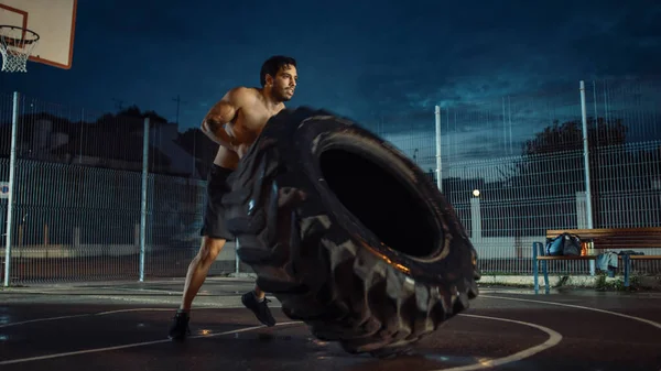 Forte Muscular Fit Young Shirtless Man está fazendo exercícios em uma quadra de basquete ao ar livre cercada. Ele está lançando um pneu pesado grande em uma tarde depois da chuva em uma área residencial de vizinhança. — Fotografia de Stock