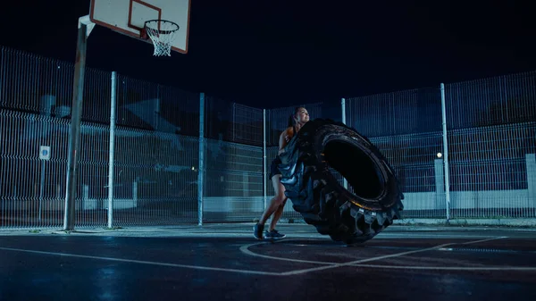Bella Energetic Fitness Girl sta facendo esercizi in un campo da basket all'aperto recintato. Shes Flipping un grande pneumatico pesante in una notte nebbiosa dopo la pioggia in una zona residenziale Quartiere. — Foto Stock