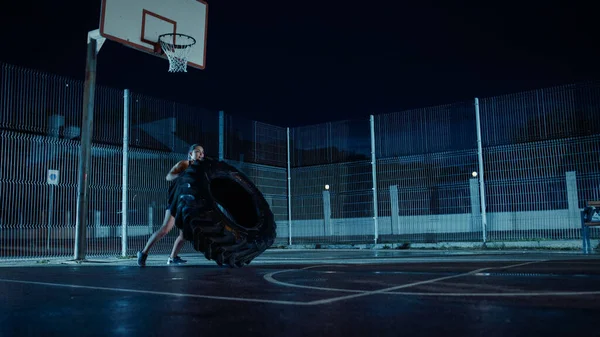 Beautiful Energetic Fitness Girl está haciendo ejercicios en una cancha de baloncesto al aire libre vallada. Ella está volteando un neumático pesado grande en una noche brumosa después de la lluvia en un barrio residencial. —  Fotos de Stock