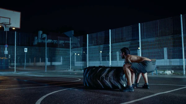 Forte forma muscolare Young Shirtless Man sta facendo esercizi in un campo da basket all'aperto recintato. Sta girando un grosso pneumatico pesante in una notte nebbiosa dopo la pioggia in una zona residenziale. — Foto Stock