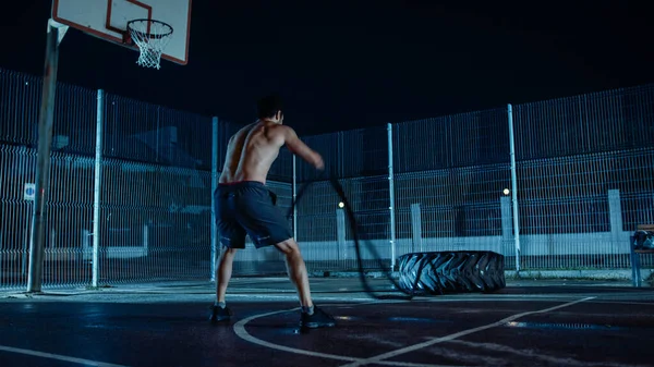 Backshot van een sterke gespierde pasvorm Shirtless Young Man doet oefeningen met Battle Ropes. Hij doet een workout in een omheind Outdoor Basketbalveld. Nachtbeelden na regen in een residentiële — Stockfoto