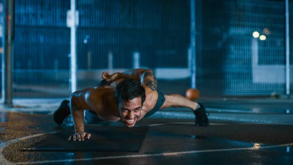 Forte Muscular Fit Shirtless Young Man está fazendo exercícios de uma mão para cima. Ele está fazendo um treino em um campo de basquete ao ar livre cercado. Noite após a chuva em uma área de vizinhança residencial. — Fotografia de Stock