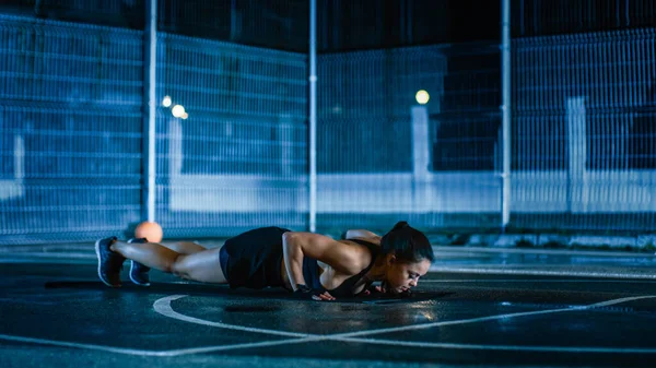 Bela aptidão desportiva menina fazendo exercícios push-up. Ela está fazendo um treino em uma quadra de basquete ao ar livre cercada. Filmagem noturna após a chuva em uma área de vizinhança residencial. — Fotografia de Stock