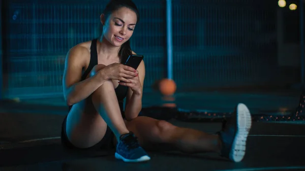 Piękna Sportowa dziewczyna fitness w czarnym stroju sportowym uśmiecha się podczas korzystania ze smartfona w nocy. — Zdjęcie stockowe