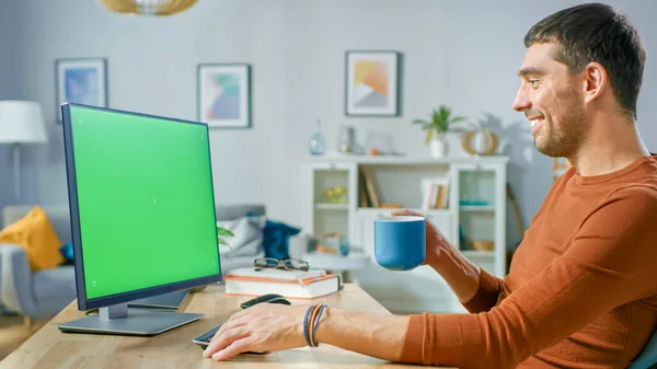 Homem Bonito Sentado em Sua Mesa em Casa Usa Computador Pessoal com Tela Verde Falsa. Ele bebe bebida da caneca. — Fotografia de Stock