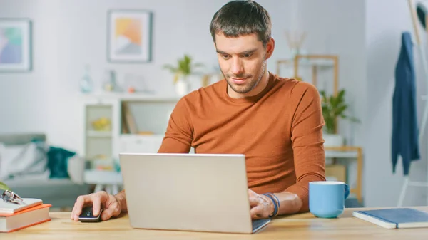 Laptopta çalışan, evdeki ahşap masasında oturan yakışıklı gülümseyen adamın portresi. İnternette gezinen adam oturma odasından defter üzerinde çalışıyor.. — Stok fotoğraf