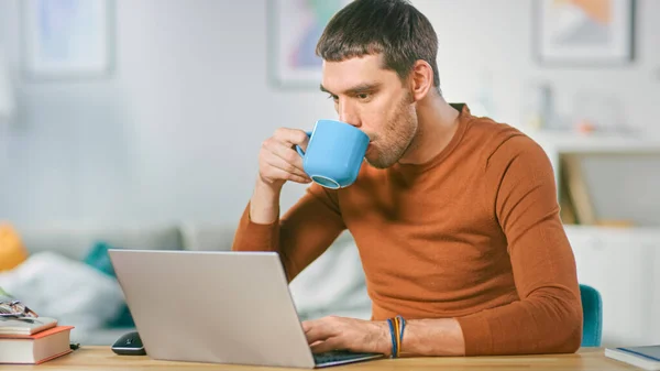 Πορτρέτο του όμορφου χαμογελαστού ανθρώπου που εργάζεται σε φορητό υπολογιστή, κάθεται στο ξύλινο γραφείο του στο σπίτι και πίνοντας καφέ. Ο άνθρωπος που περιηγείται στο Διαδίκτυο, που εργάζονται σε σημειωματάριο από το σαλόνι του. — Φωτογραφία Αρχείου