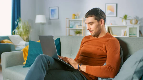 Komea mies käyttää kannettavaa tietokonetta istuessaan sohvalla kotona. Mies työskentelee, selailee Internetiä kodikas olohuone. — kuvapankkivalokuva