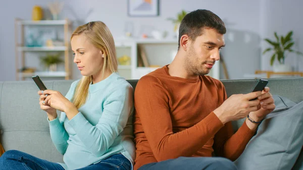 Konzept des Entfremdungsstreits: Zu Hause auf dem Sofa sitzt ein junges Paar getrennt, stellt sich unterschiedlichen Richtungen und nutzt Smartphones. — Stockfoto