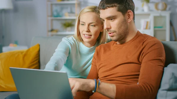 Bonito casal jovem senta-se em um sofá em casa, Eles usam computador portátil e se divertir. Retrato de belo casal jovem feliz no amor. — Fotografia de Stock