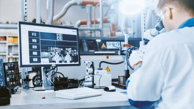 Beyaz İş Ceketi Giyen Elektronik Fabrikası İşçisi, Dijital Mikroskoba bağlı bir Bilgisayar Ekranındaki Yazılı Devre Tahtasını Denetliyor. Yüksek Teknoloji Fabrikası.