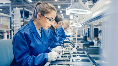 Mavi İş Ceketi ve Koruyucu Gözlüklü Kadın Elektronik Fabrikası İşçisi Akıllı telefonları tornavidayla monte ediyor. Arka planda daha fazla çalışanı olan Yüksek Teknoloji Fabrikası.