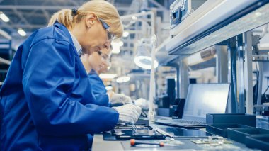 Mavi İş Ceketi ve Koruyucu Gözlüklü Kadın Elektronik Fabrikası İşçisi, Akıllı Telefonları Tornavidayla Birleştiriyor. Arka planda daha fazla çalışanı olan Yüksek Teknoloji Fabrikası.